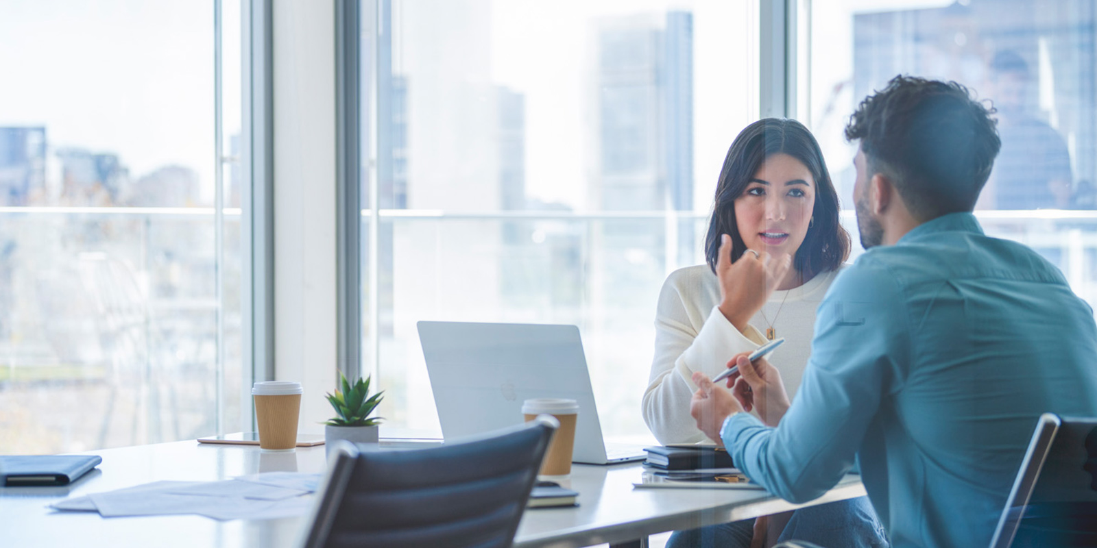 Two people talking at a table in an office with blurred city view in the background.