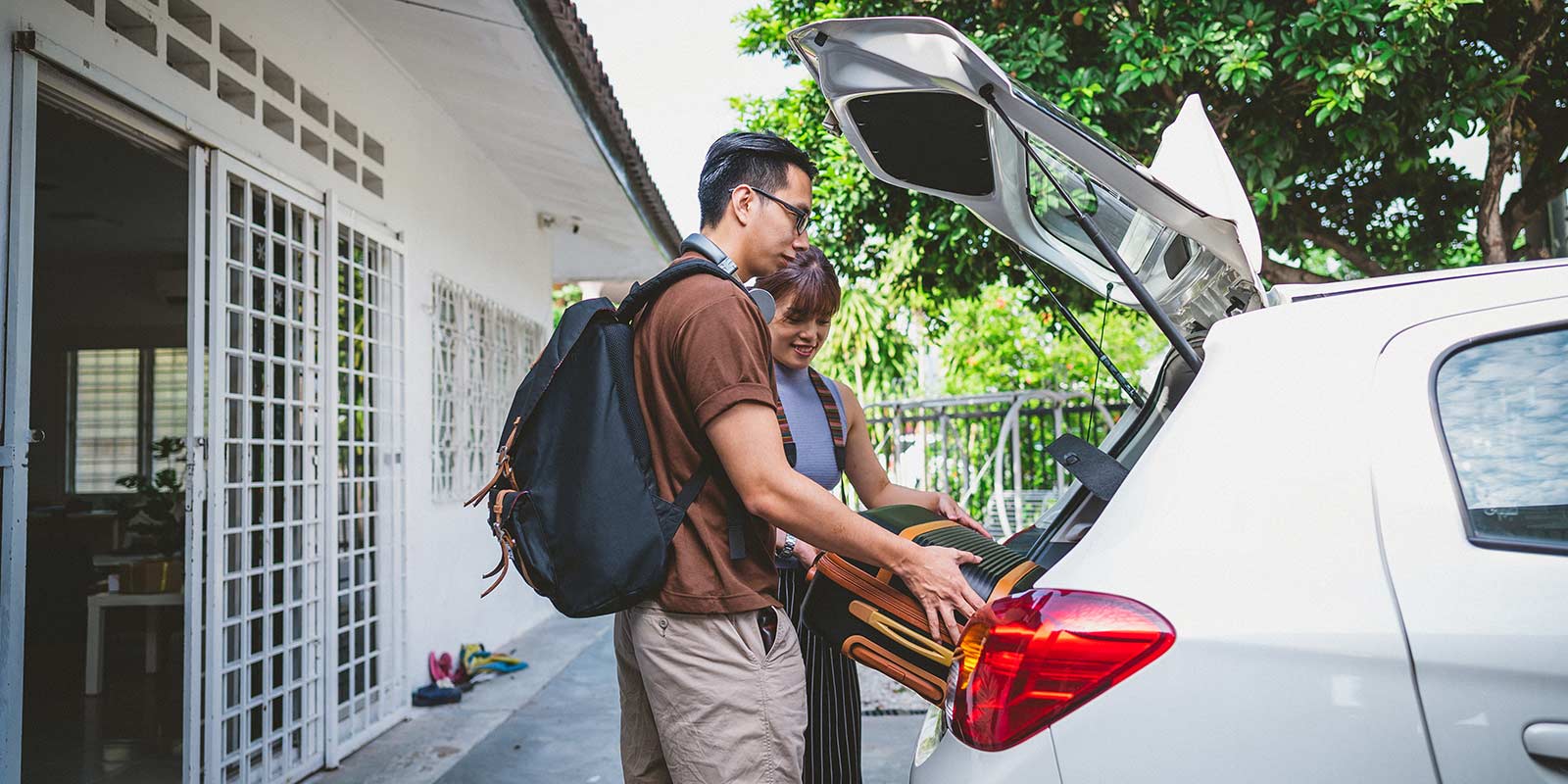Two people loading a suitcase into the open boot of a white car outside a house.