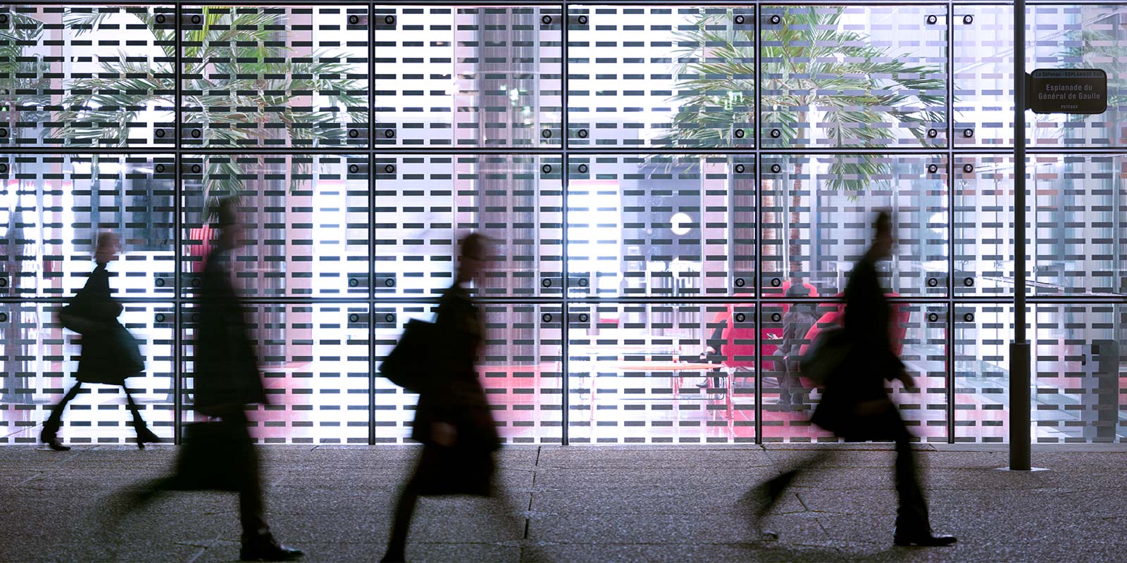 Urban scene with blurred figures walking past a large glass wall with indoor plants and furniture visible behind it; signpost on the right.