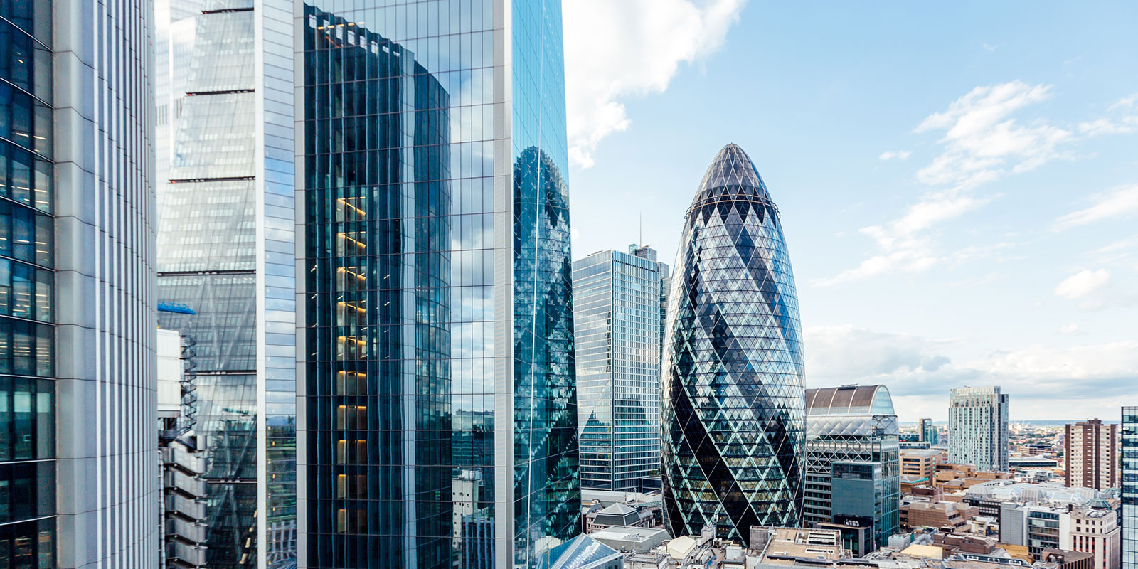 Cityscape with modern skyscrapers, including the Gherkin, surrounded by reflective glass buildings under a partly cloudy sky.