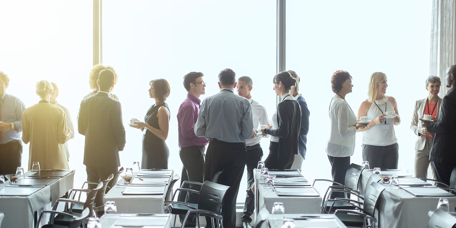 Group of people conversing in a conference room with rows of tables and chairs, some set with glasses and notepads, and large windows in the background.