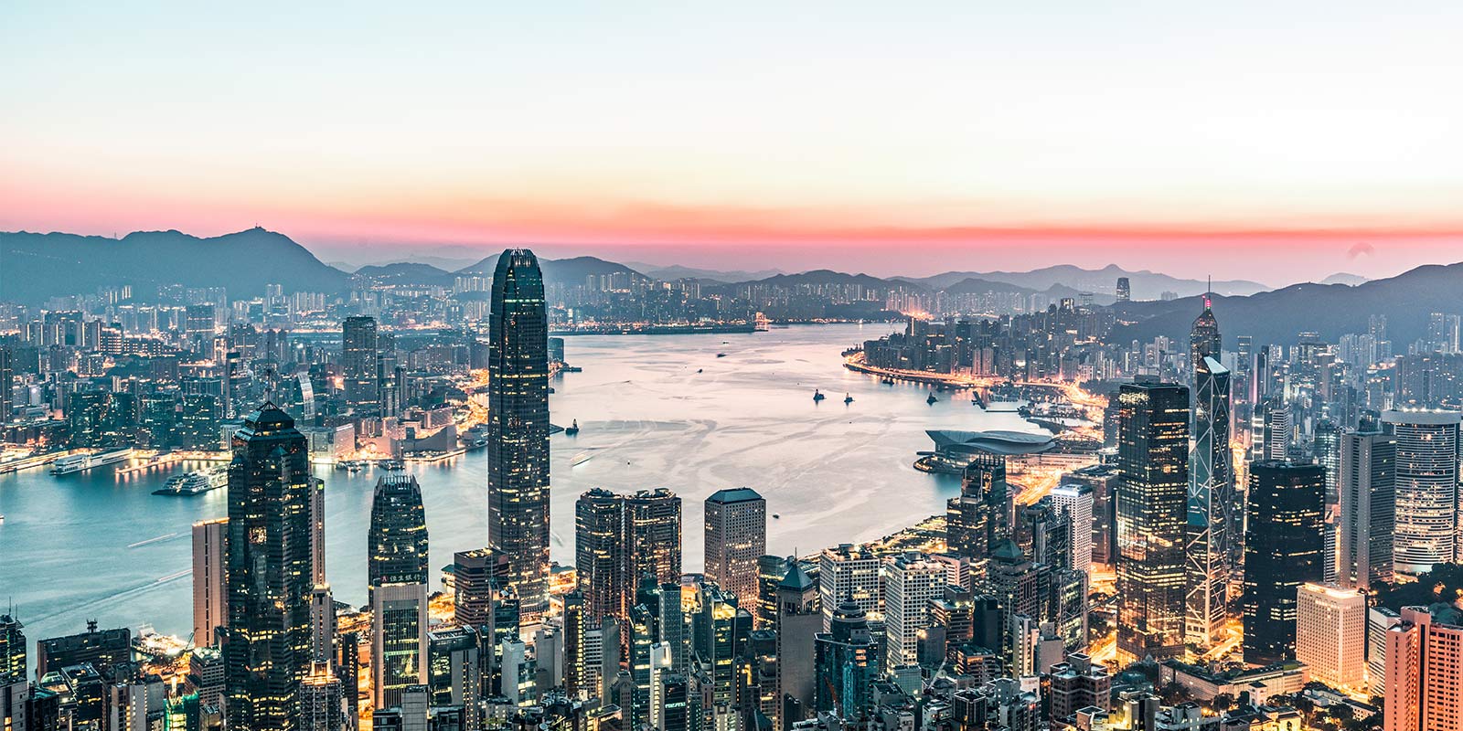 Hong Kong skyline at dusk with skyscrapers, Victoria Harbour, and mountains under an orange-blue sky.