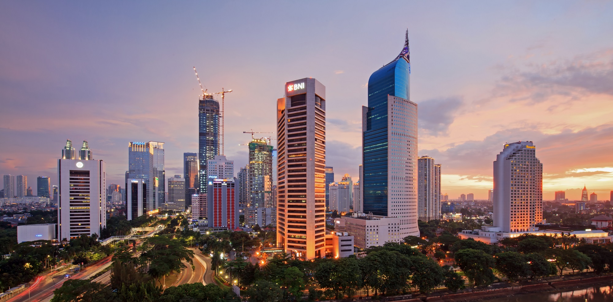 Jakarta skyline at sunset with tall buildings, cranes, and orange-pink sky.