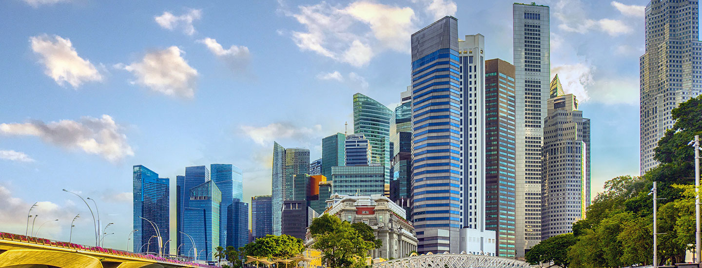 Singapore cityscape featuring the Fullerton Building in the foreground, modern skyscrapers, a bridge on the left, and greenery under a blue sky with scattered clouds.