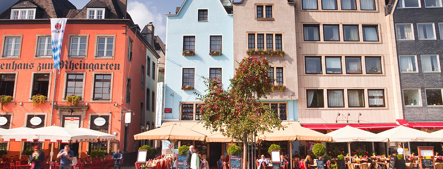 Street view in Cologne with colorful buildings, including Brauhaus zum Rheingarten, outdoor café seating, and a tree with red berries.