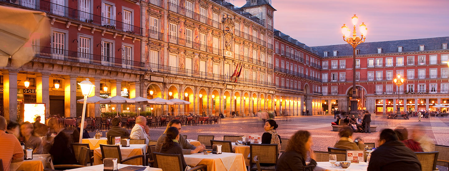 Outdoor plaza at sunset with people dining at café tables, surrounded by an ornate building with balconies and lit street lamps.
