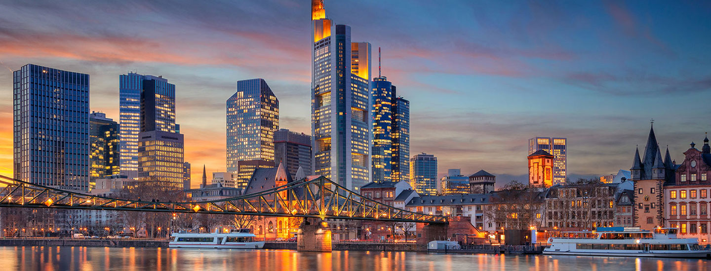 Panoramic sunset view of Frankfurt with the Commerzbank Tower, modern skyscrapers, a bridge over the Main River, and historic buildings on the right.