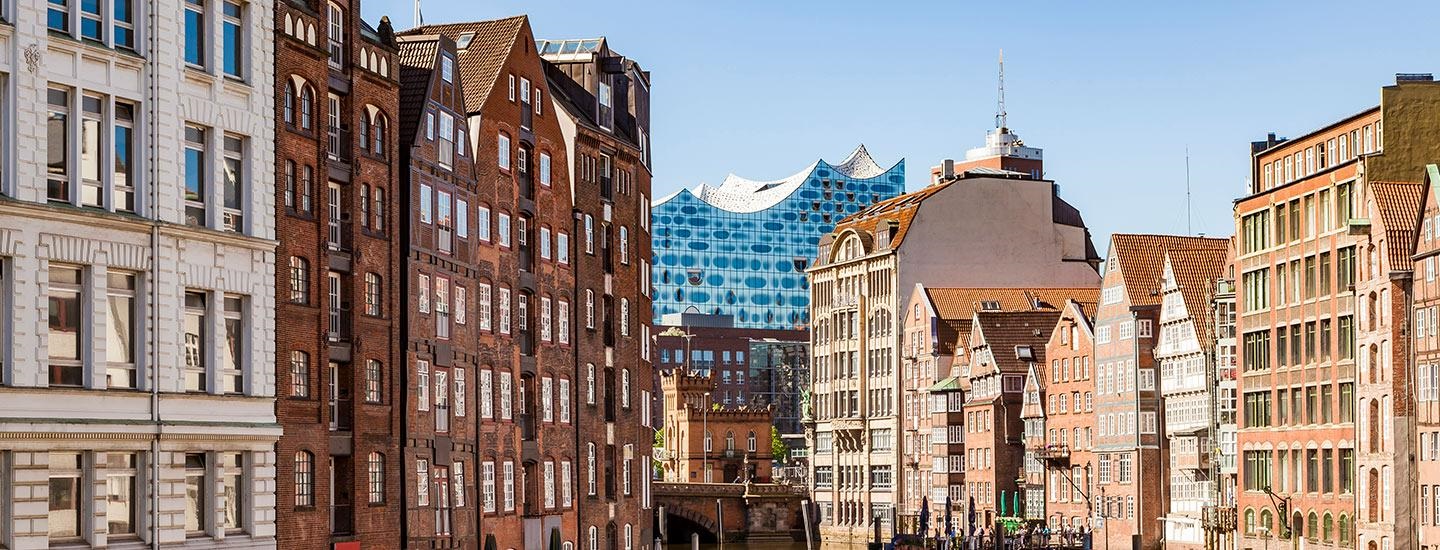 Hamburg cityscape with traditional brick buildings in the foreground and the Elbphilharmonie’s wavy glass roofline in the background under a clear blue sky.