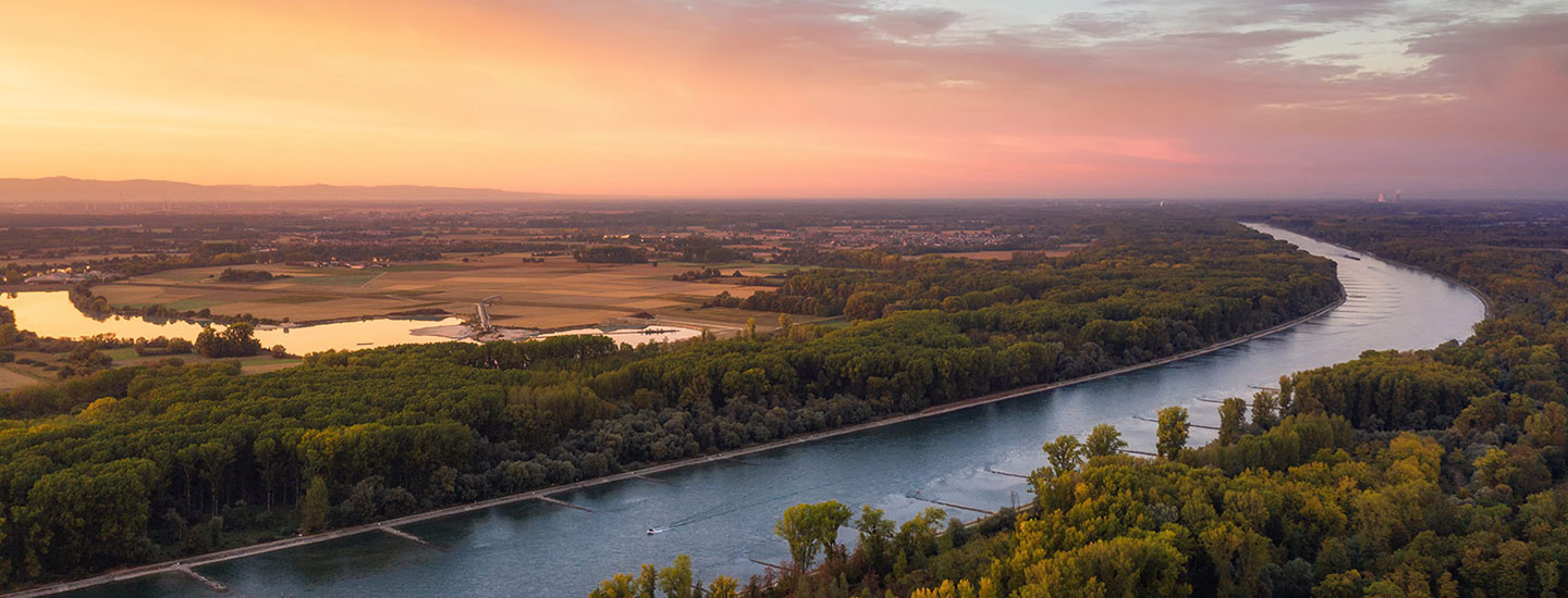 Aerial view near Karlsruhe showing a winding river through green forests and farmland under a warm orange-pink sunrise or sunset sky.