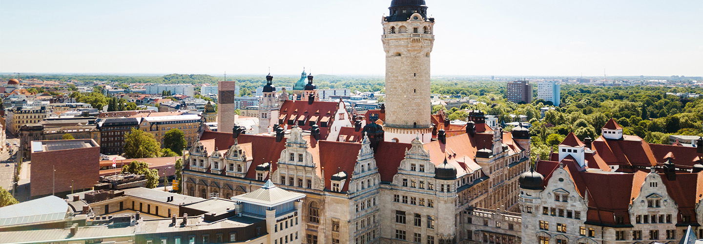 Panoramic view of Leipzig featuring the New Town Hall with its tall tower and red-tiled roofs, surrounded by historic buildings and greenery under a clear blue sky.