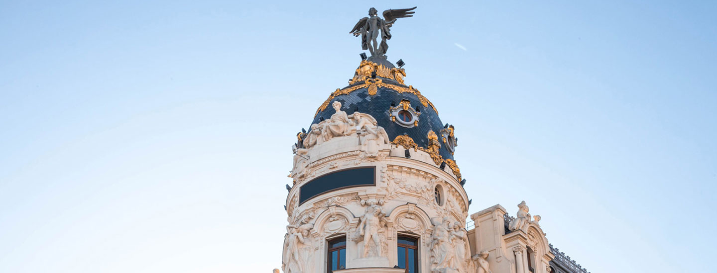 Top of the Metropolis Building in Madrid with a dark blue dome, golden details, and a winged statue, surrounded by ornate sculptures.