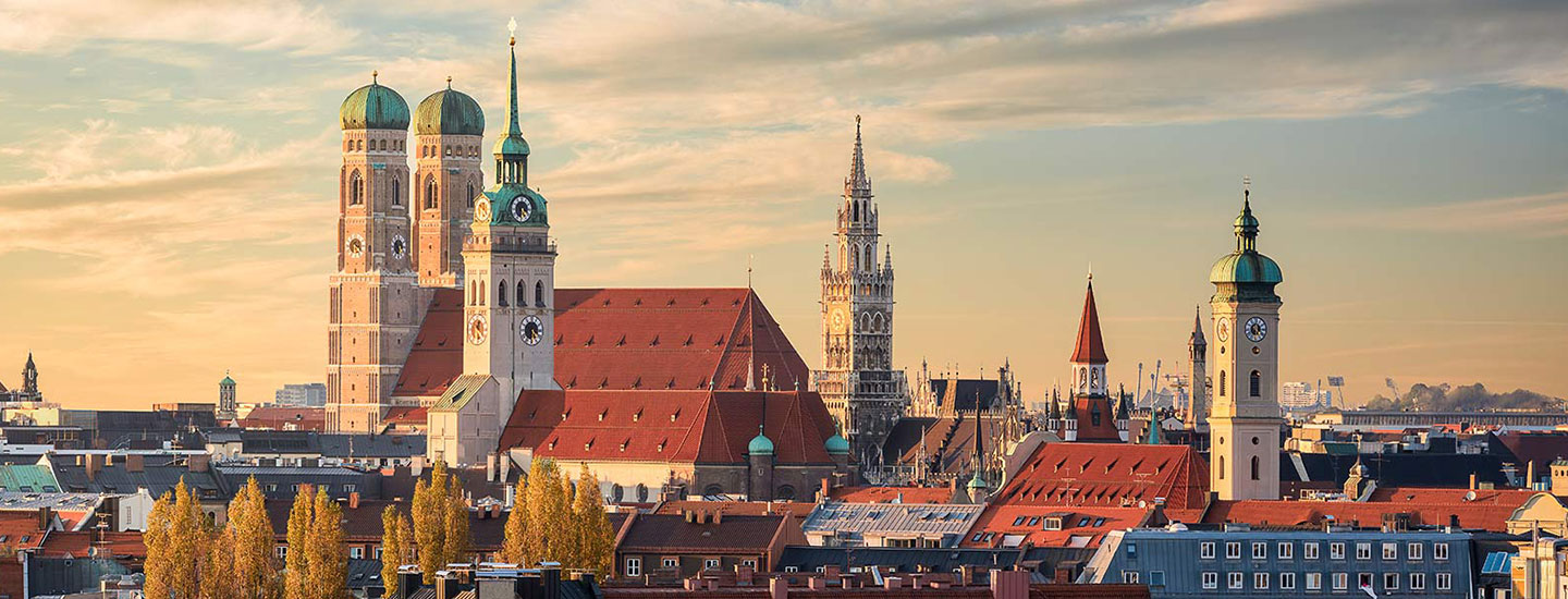 Panoramic view of Munich with the Frauenkirche, New Town Hall, and St. Peter's Church under a partly cloudy sky at sunset.
