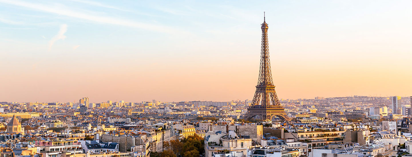 Panoramic view of Paris with the Eiffel Tower centered, surrounded by city buildings under a warm sunset sky.