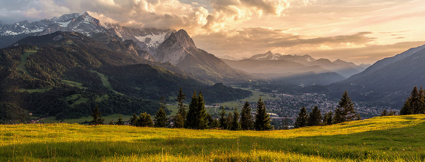 Scenic German landscape with green meadows, scattered trees, snow-capped mountains, and a small town in a valley at sunrise or sunset.