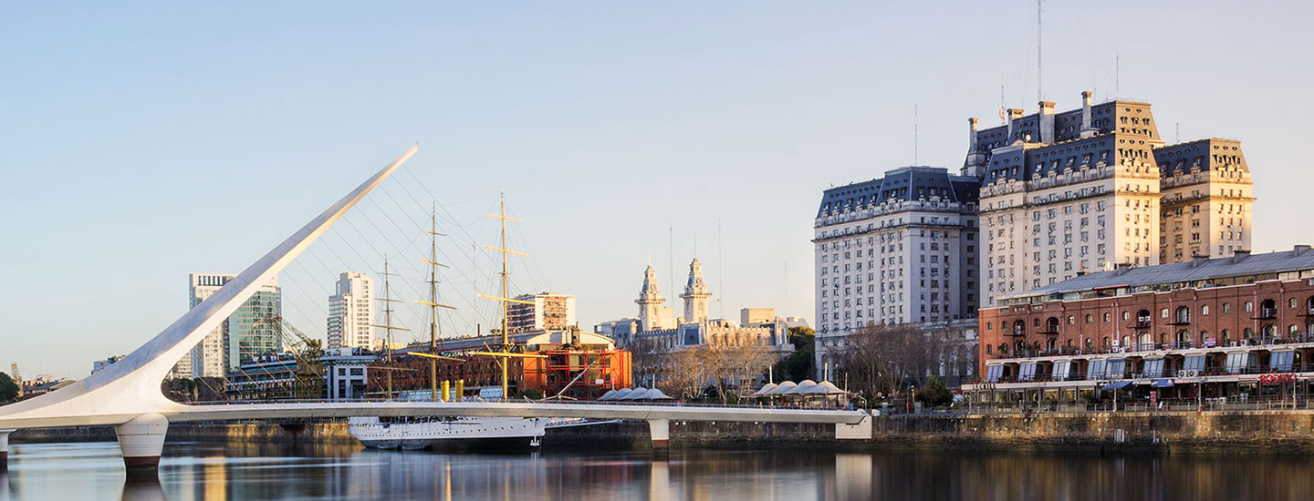 Cityscape in Buenos Aires, Argentina, featuring Puente de la Mujer bridge over water, a docked sailing ship, and historic buildings.