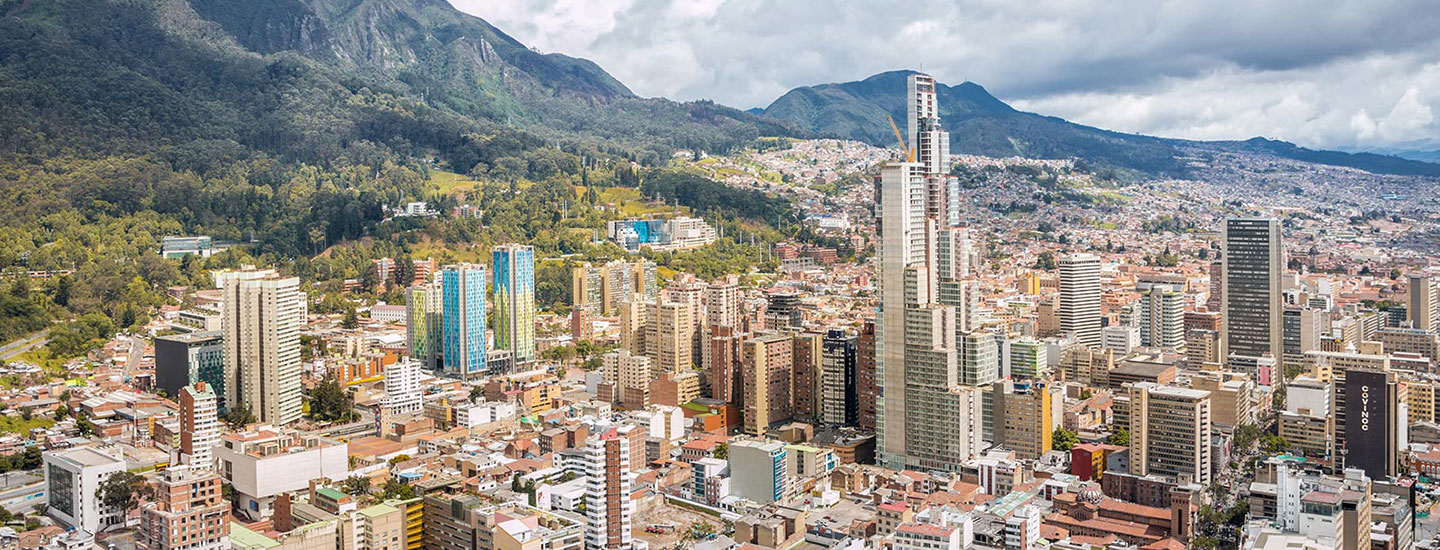 Panoramic view of Bogotá D.C., Colombia, with high-rise buildings and lush green mountains in the background under a cloudy sky.