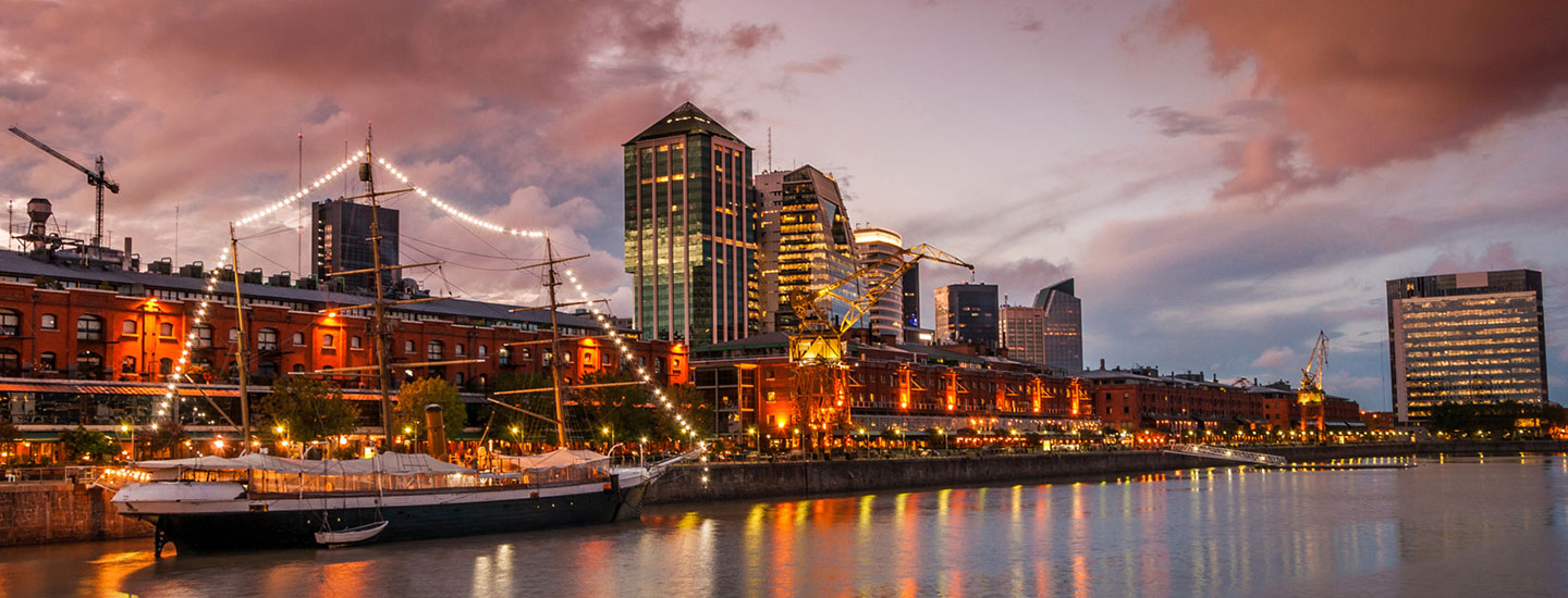 Evening view of Buenos Aires waterfront with a lit docked ship, modern high-rises, historic brick buildings, and a colorful sky reflected on calm water.