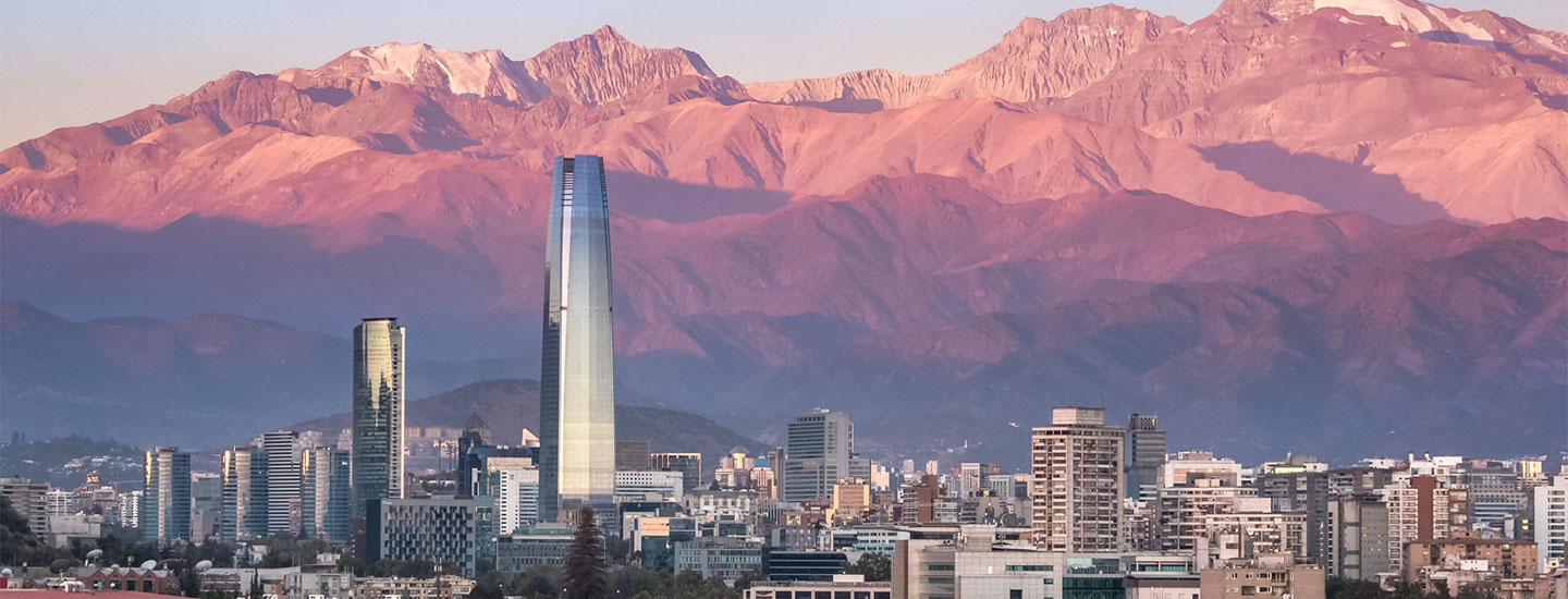 City skyline in Chile with modern buildings and a mountain range in the background, bathed in pinkish sunset light.