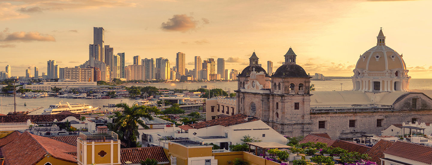 Panoramic view of Cartagena, Colombia at sunset, featuring historic red-tiled buildings and a domed church in the foreground, with modern high-rises along the coastline.