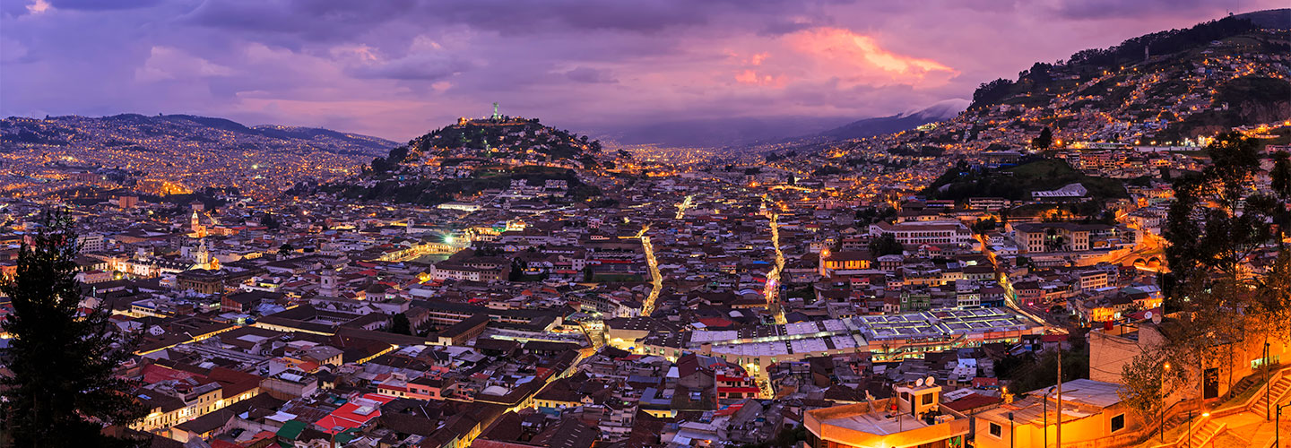 Twilight panoramic view of Quito, Ecuador, with city lights glowing against surrounding hills and mountains, and La Virgen de El Panecillo statue visible in the distance.