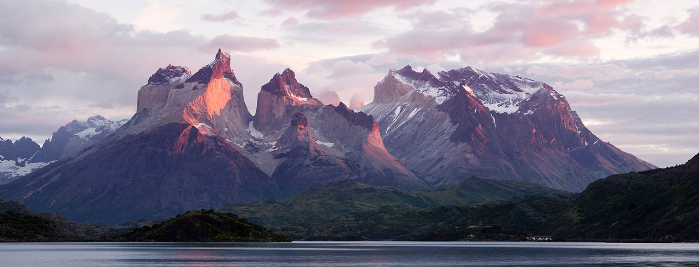 Scenic view of Torres del Paine in Patagonia, Chile, with rugged peaks, snow patches, and a reflective body of water under a pink and purple sunrise or sunset sky.