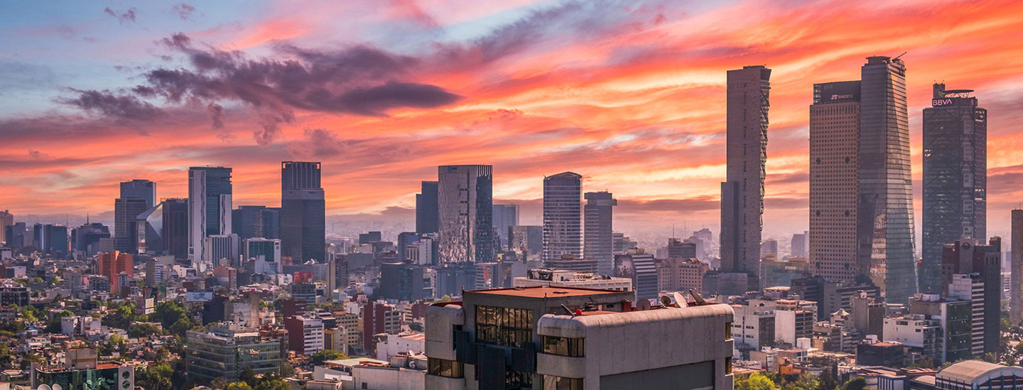 Panoramic sunset view of Mexico City with high-rise buildings including Torre Reforma and BBVA Bancomer Tower, set against a vibrant orange, pink, and purple sky.