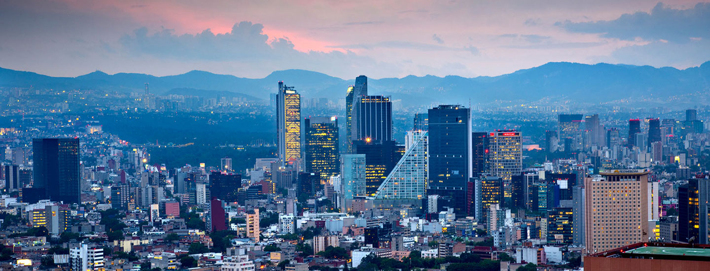 Panoramic view of Mexico City at dusk, with a dense skyline of modern and historic buildings, set against mountains and a cloud-streaked sunset sky.