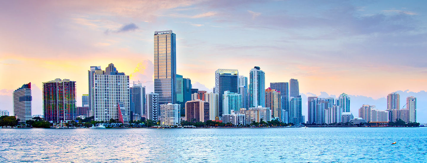 Skyline of Miami, Florida with waterfront high-rises under a partly cloudy sky, reflecting warm sunrise or sunset colors in the water.