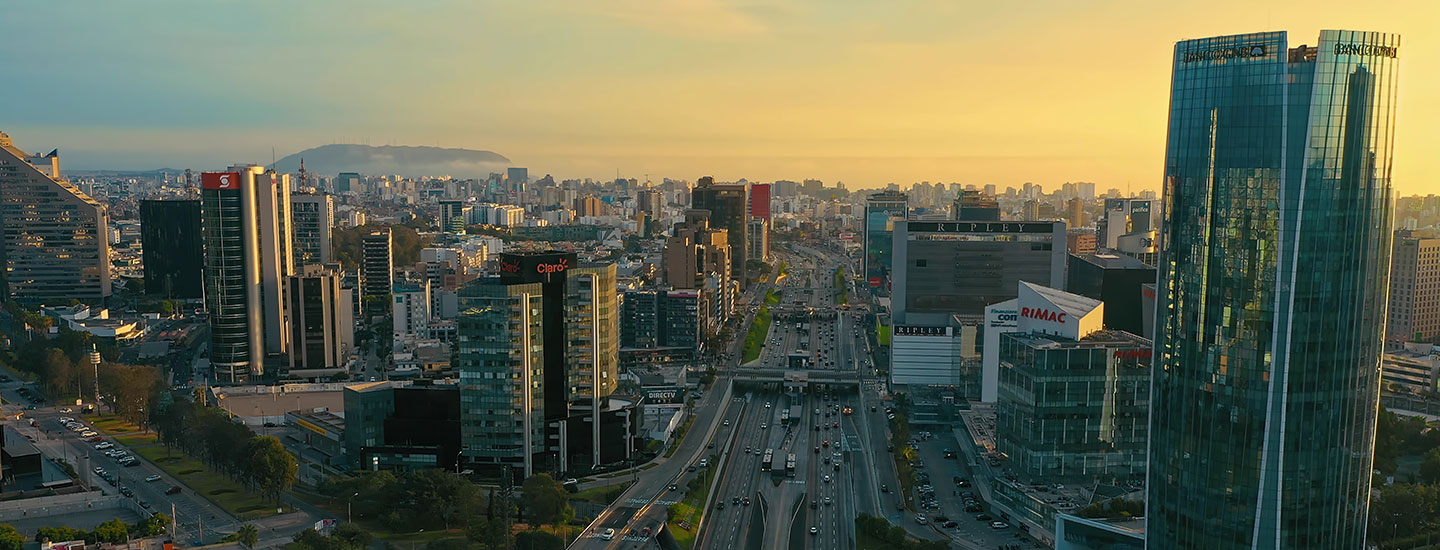 Panoramic sunset view of Lima, Peru, featuring modern high-rises like Interbank Tower, a busy central highway, and distant hills under a golden sky.