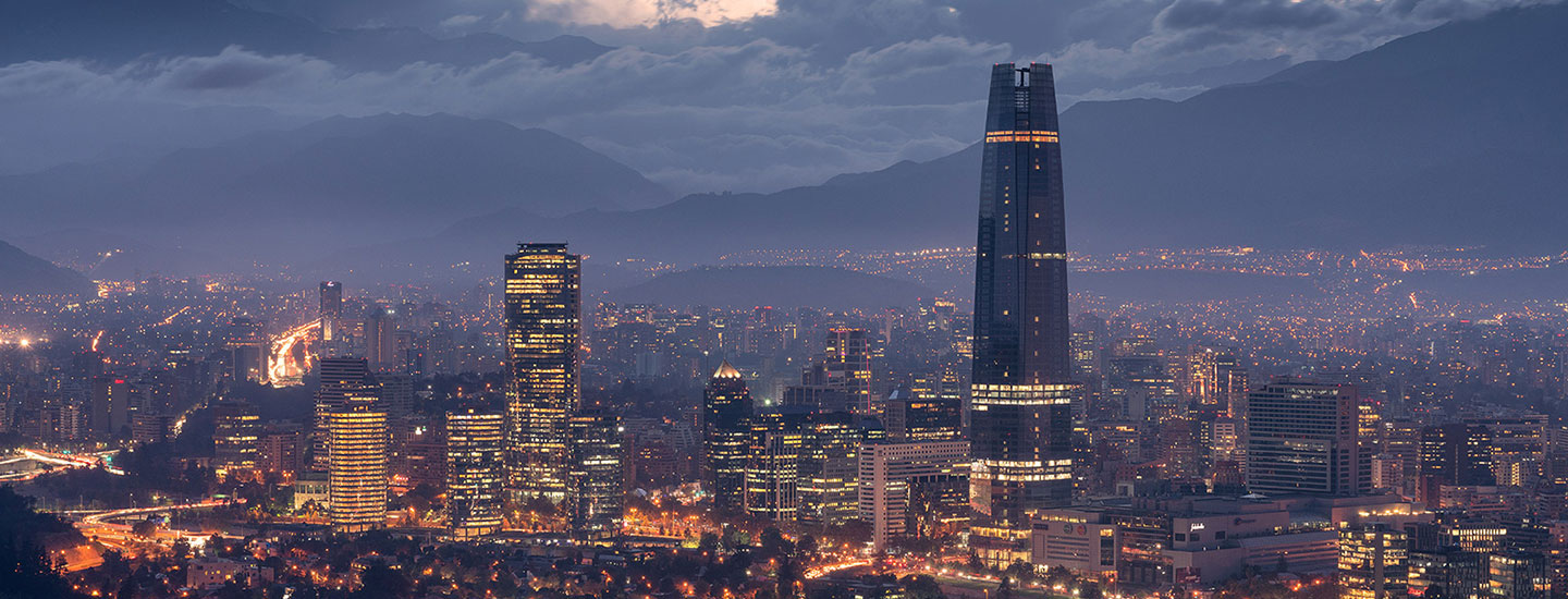 Nighttime cityscape of Santiago, Chile, featuring the illuminated Gran Torre Santiago and surrounding high-rises, with mountains in the background under a cloudy sky.