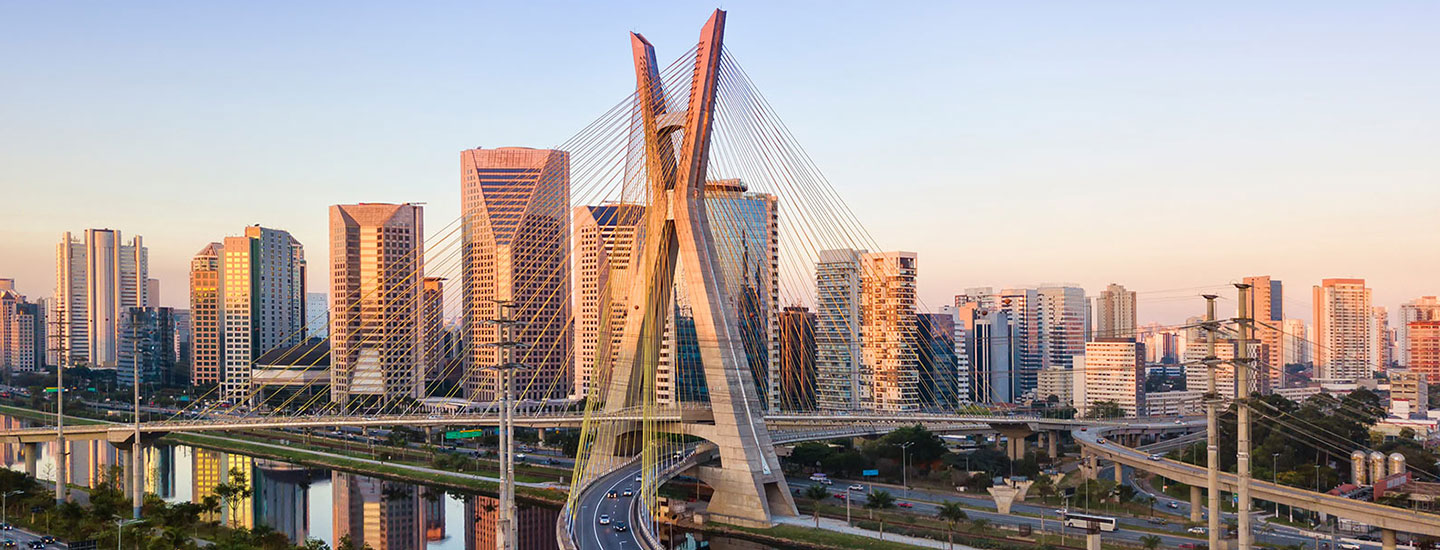 Cityscape of São Paulo, Brazil featuring a cable-stayed bridge with curved pylons, modern high-rises, and a river, captured in warm early morning or late afternoon light.