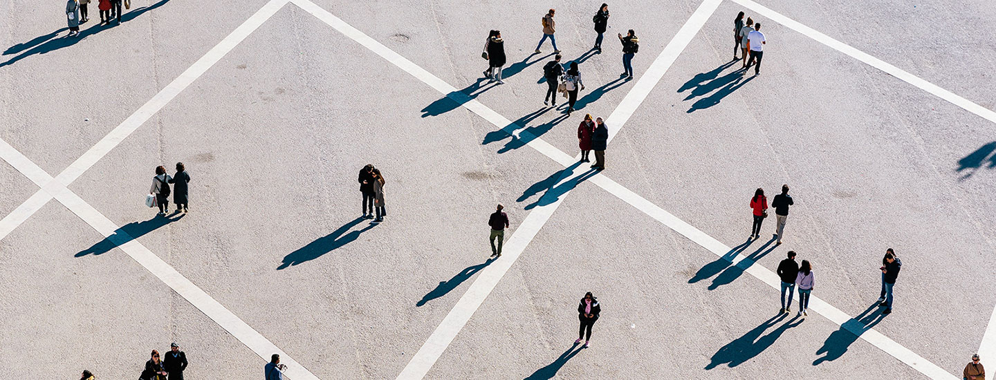 Aerial view of a geometric open space with intersecting white lines and scattered groups of people casting long shadows, likely taken during sunrise or sunset. City unspecified.
