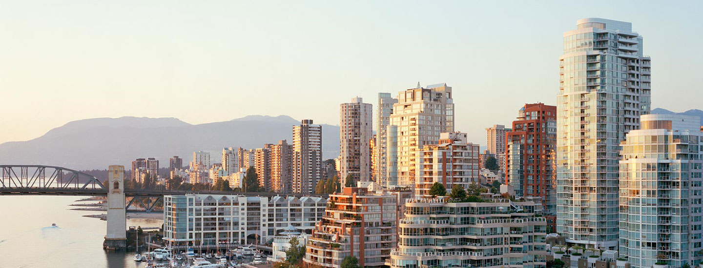 Cityscape of Vancouver, Canada with modern high-rises, a bridge over a marina with docked boats, and mountains in the background under a clear sky.