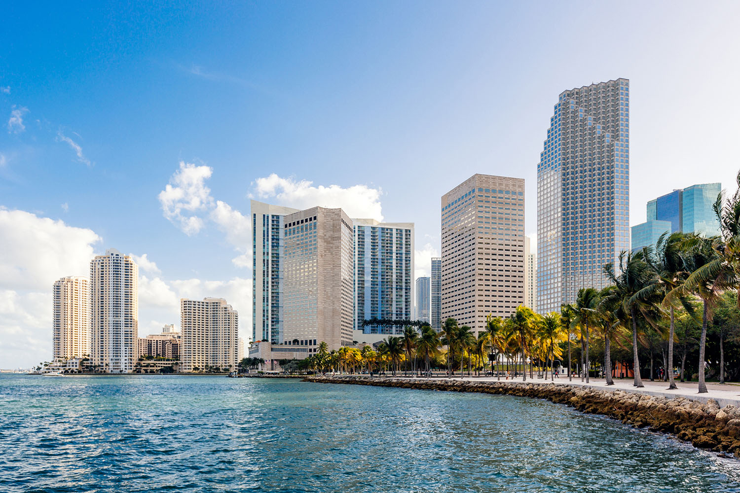 Waterfront cityscape with modern buildings, palm-lined promenade, and scattered clouds.
