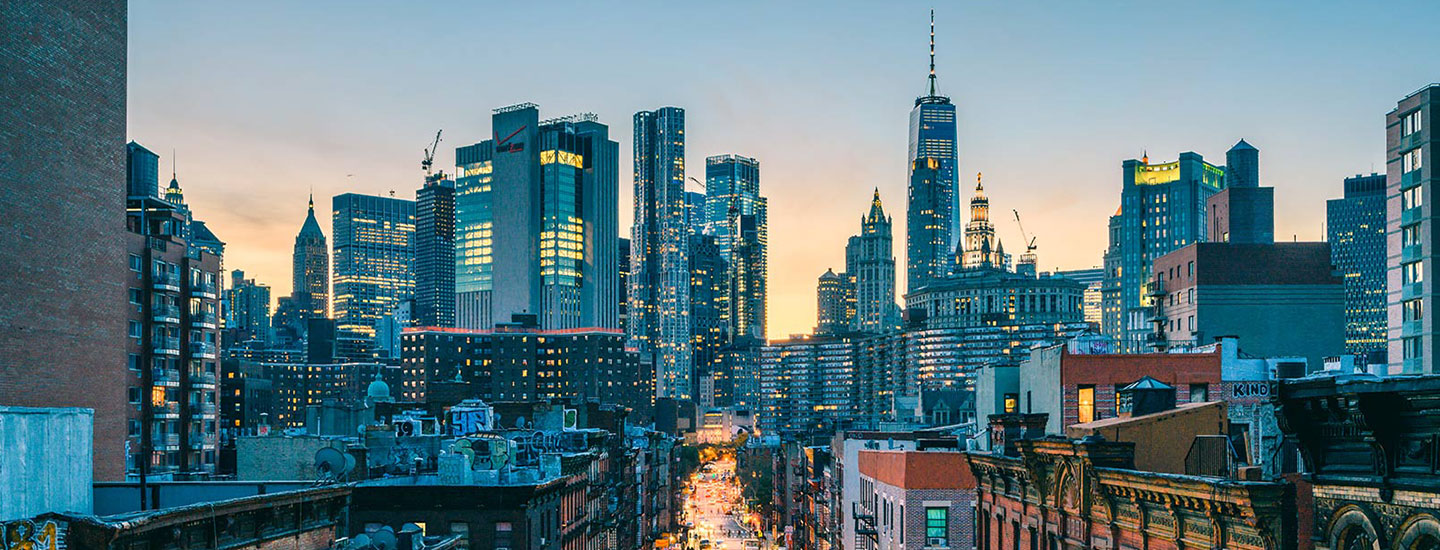 Dusk cityscape of New York City with illuminated skyscrapers under a gradient sky transitioning from light blue to orange.