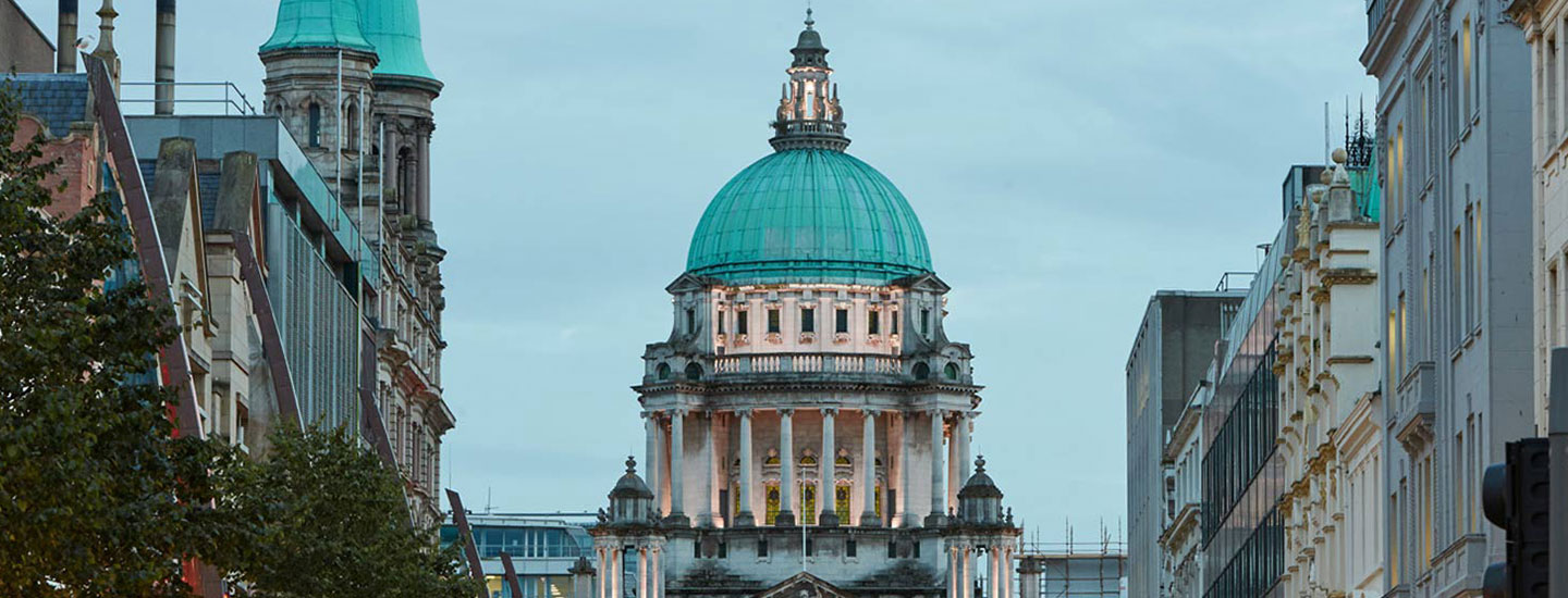 Street view in Belfast, Northern Ireland, featuring a prominent historical building with a green dome at the center, surrounded by urban architecture.