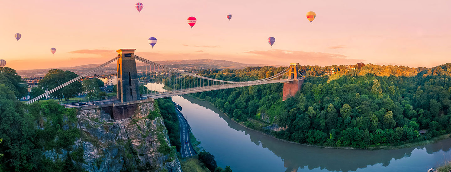 View of the Clifton Suspension Bridge in Bristol, England, spanning the Avon Gorge with hot air balloons in the sky and a cityscape in the background during sunrise or sunset.