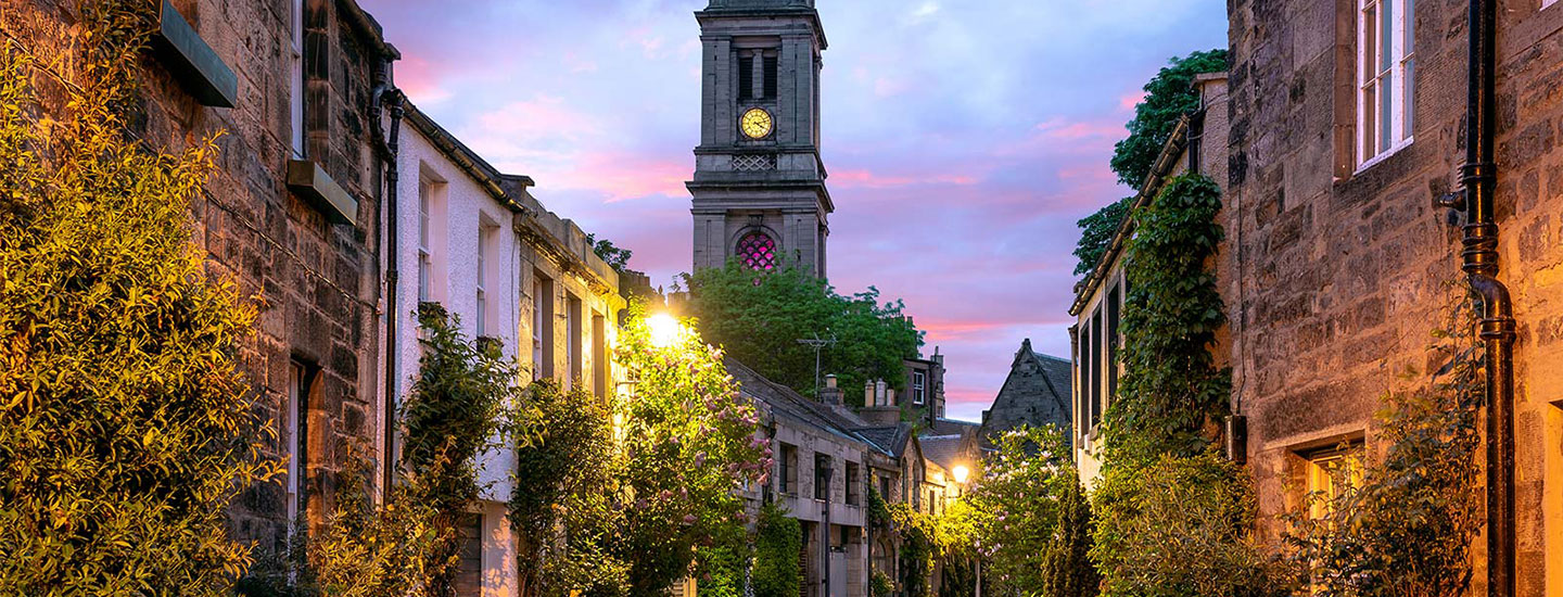 Sunset view of a picturesque street in Edinburgh, Scotland, with old stone buildings, warm streetlights, and a prominent clock tower under a pink and purple sky.