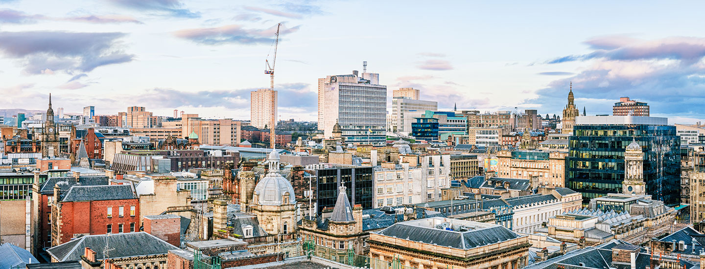 Panoramic view of Glasgow, Scotland, featuring a mix of historic and modern architecture, including domes, spires, and high-rises under a partly cloudy sky.