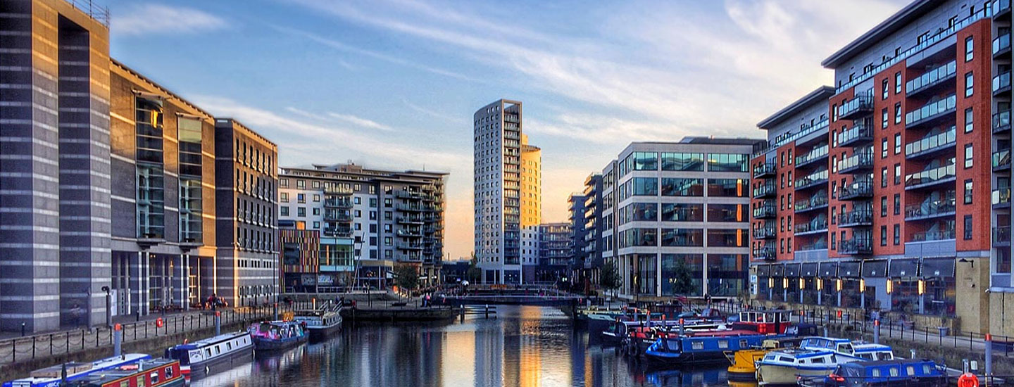 Modern canal scene in Leeds, England, with docked boats, contemporary buildings in varied styles, and a tall structure in the background under a clear sky.
