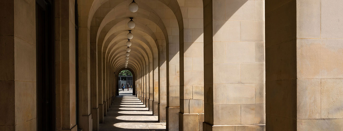 Arched stone corridor in Manchester, England, with evenly spaced columns, hanging spherical lights, and sunlight casting shadows along the walkway.