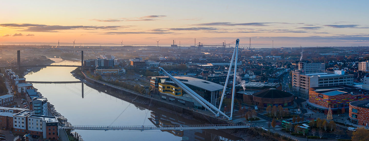 Panoramic view of Newport, Wales at sunrise with the River Usk, city footbridge, and distant wind turbines.