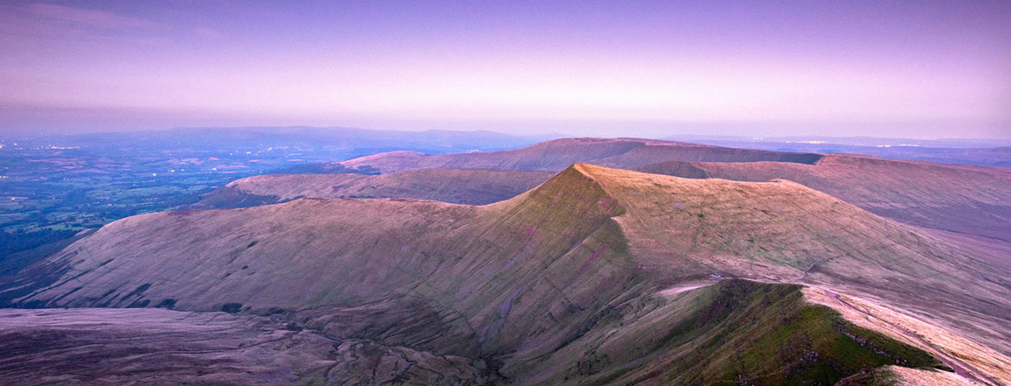 Sunset over the Brecon Beacons in Wales, showing rolling green and brown hills under a fading blue-purple sky.