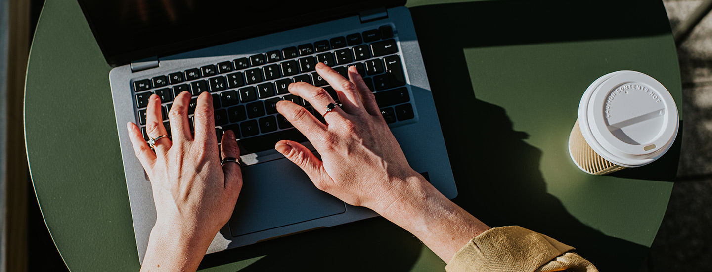 Person typing on a laptop placed on a green surface, with a disposable coffee cup beside it.