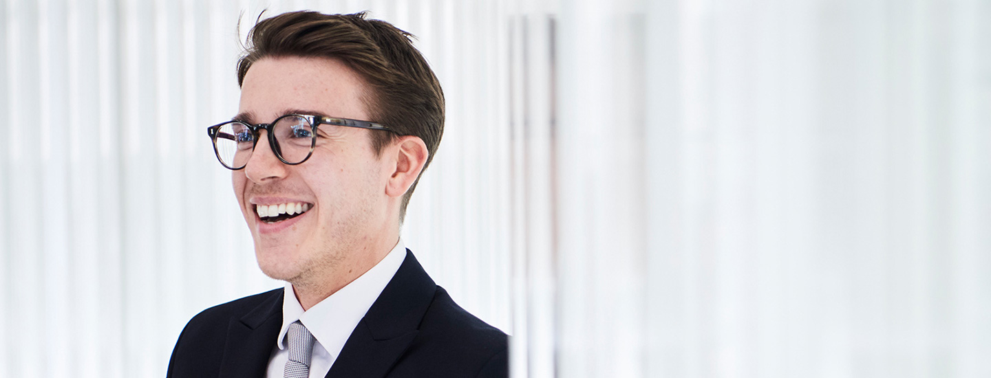 Person in a dark suit with a white shirt and light tie standing in front of vertical white blinds, face blurred