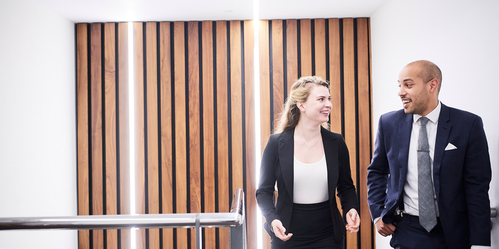 Two people in business attire walking past a wall with vertical wooden slats and white lighting