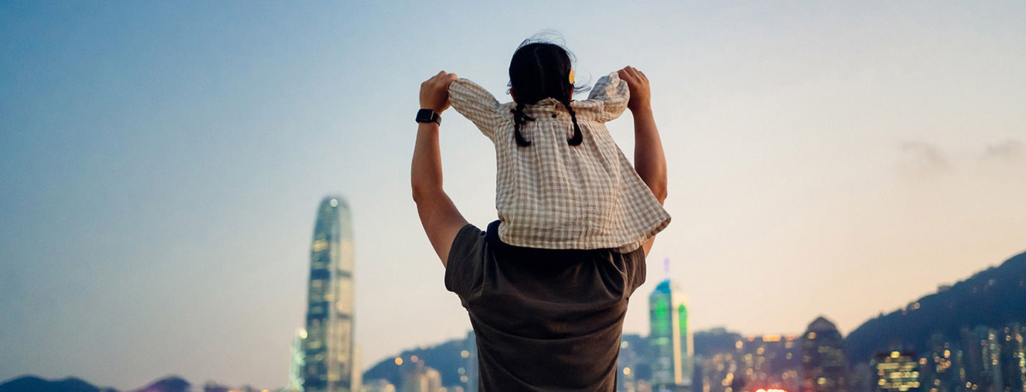 Person carrying a child on their shoulders, facing a cityscape with tall buildings and mountains at dusk or dawn