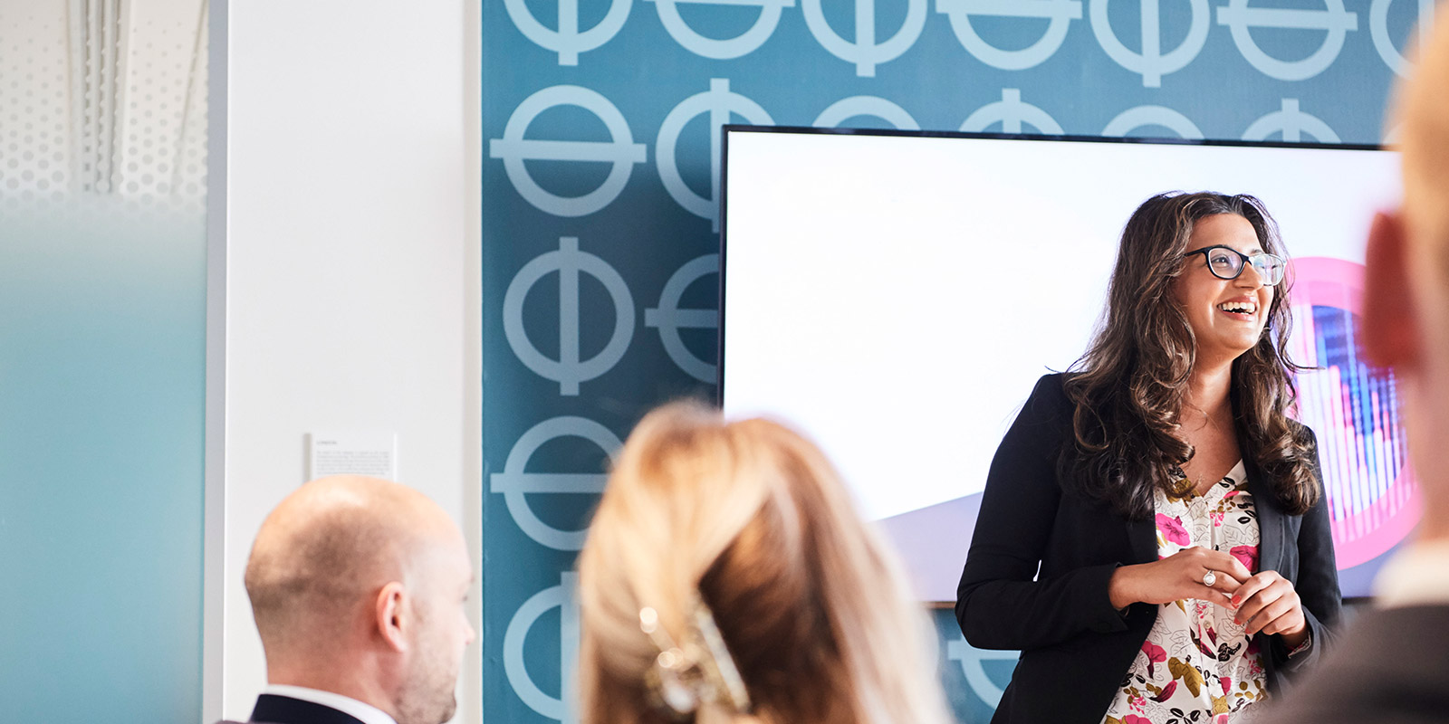 A person presenting in front of a group with a screen and patterned blue wall in the background.