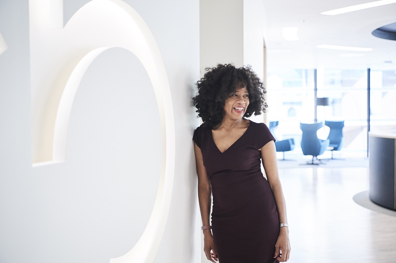  Person with curly hair in a dark dress leaning against a wall with an illuminated letter "b"; modern office setting with large windows.