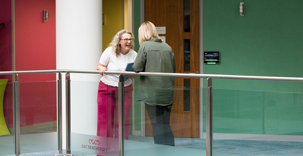 Two individuals conversing in a hallway with colourful walls, a white column, wooden door, and glass railing displaying the text 